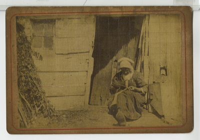 390103 Vrouw met naaiwwerk zittend in deuropening van een houten huis, ca. 1890