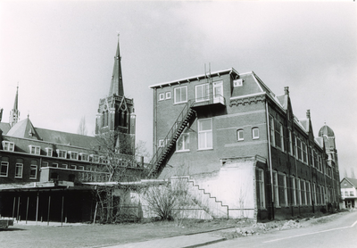 240276 Schoolpad: achtergevel Heilig Hartschool met op achtergrond de toren van de Sint Odulphuskerk, 1985