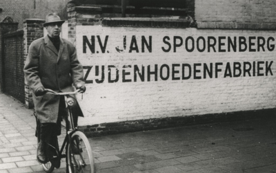 4267 Jan Spoorenberg: directeur van N.V. Jan Spoorenberg Zijdehoedenfabriek, op de fiets naast een muur met opschrift ...