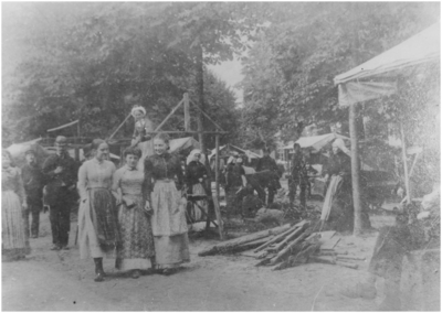 21753 Drie dames wandelend over de Markt, waar markt- of kermiskramen worden opgebouwd. In het midden juffr. Egelie, ca. 1900