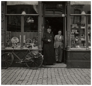 188039 De heer Muffels samen met een geestelijke voor zijn tabaks- en sigarenwinkel, ca. 1940