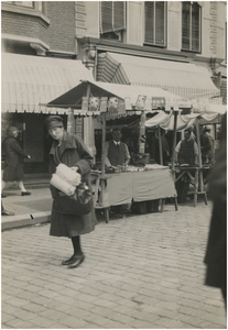 192946 Het doen van boodschappen. Op de achtergrond een marktkraam met scheergerei, ca. 1930