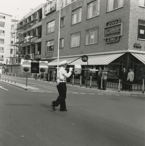 195745 Politieman met stoplicht op zijn schouder steekt de weg over, 1961-1965