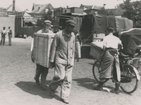195698 Het versjouwen van een kist door twee mannen, passerende man met vrachtfiets, op de achtergrond vrachtkarren, 06-1940
