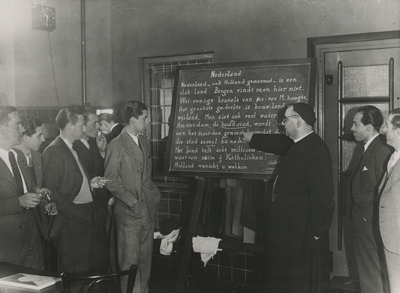 195854 Priester (onderwijst de Nederlandse taal) wijst op een schoolbord naar een woord aandachtig gevolgd door de ...