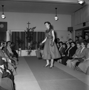 255024 Mannequin showt toeschouwers een jurk op de catwalk, 1955