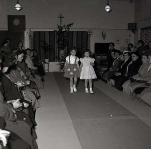 255030 Twee kleuters meisjes tonen de toeschouwers hun jurkjes op de catwalk, 1955