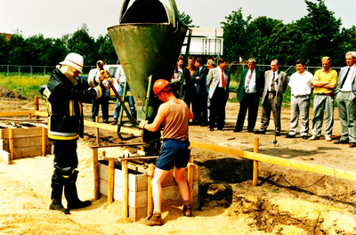 232557 Het storten van de eerste beton voor de fundering van de nieuwbouw door burgemeester Vos. Aan de rechterkant van ...