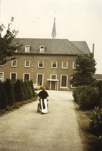 265398 Een kruisheer duwt een rolstoel over het pad naar het klooster vanuit de kloostertuin te St. Agatha