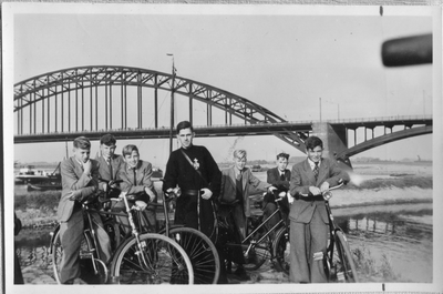 276118 Studenten van het Gabriëlcollege te Mook met een pater aan de Waal met de Waalbrug op de achtergrond in Nijmegen