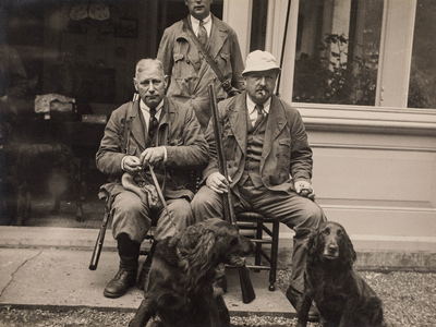  Drie jagers, waarvan twee zittend, met twee honden voor de veranda van huis Wickenburgh te 't Goy