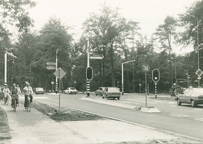 10485 - kruispunt beveiligd met verkeerslichten; mensen op fietspad; auto's op de weg; lantaarnpalen met anbw-borden