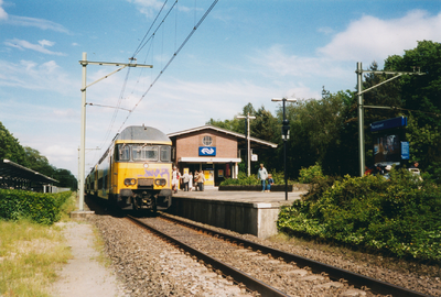 N 10111 - treinstel van NS staat aan een perron bij een stationsgebouw; links een overdekte rijwielstalling.
