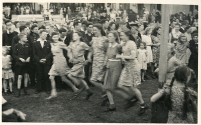 16806 - Meisjes in jurken dansen onder begeleiding van het harmonieorkest in de muziektent. De jongeman geheel links ...