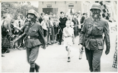 16844 - Op de voorgrond lopen twee burgers verkleed als Duitse soldaten met gasmasker op. Achter hen loopt een jongen ...