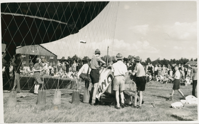 16937 - Grote groep mensen, burgers en militairen kijken naar het oplaten van een luchtballon. Padvinders met hoeden op ...
