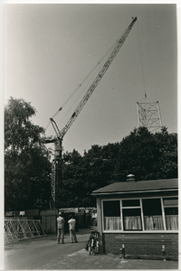 3775 - Een kraan hijst delen van een antennemast op. De mast wordt ten behoeve van de Centrale Antenne Inrichting (CAI) ...