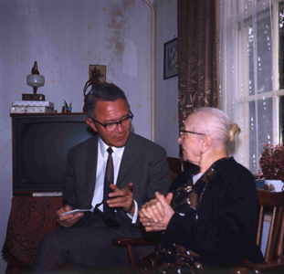 DIA_GF_1110 Een oudere vrouw (lid van de katholieke kerk) in haar huiskamer, met de pastoor; ca. 1967