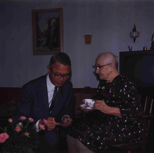 DIA_GF_1112 Een oudere vrouw (lid van de katholieke kerk) in haar huiskamer, met de pastoor; ca. 1967