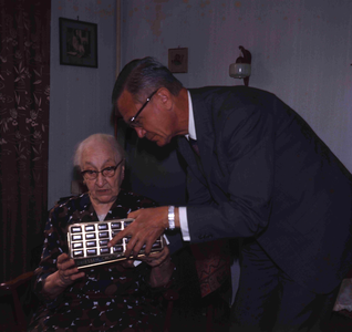 DIA_GF_1114 Een oudere vrouw (lid van de katholieke kerk) in haar huiskamer, met de pastoor; ca. 1967