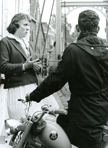 BR_TURFKADE_021 Een meisje leunt tegen de gevel van Hotel de Doelen, in gesprek met een jongen op een brommer; 1961