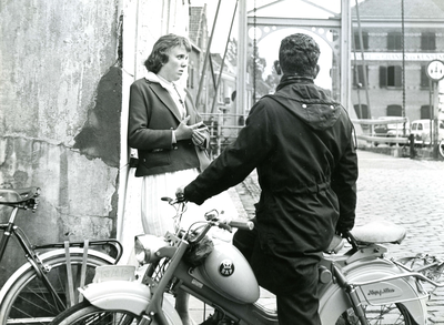 BR_TURFKADE_022 Een meisje leunt tegen de gevel van Hotel de Doelen, in gesprek met een jongen op een brommer; 1961