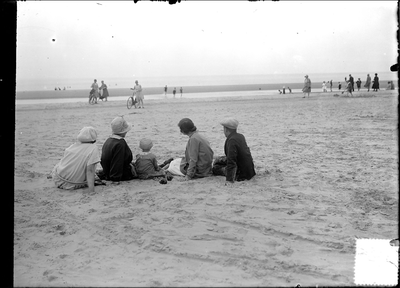 GN2362 Een familie (gezien vanaf de rug) zit op het strand en kijkt naar andere badgasten met fietsen; ca. 1925