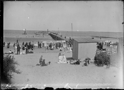 GN2674 Kijkje op het strand met badgasten en de Noordzee. Langs het aanlegsteiger ligt de veerboot; ca. 1925