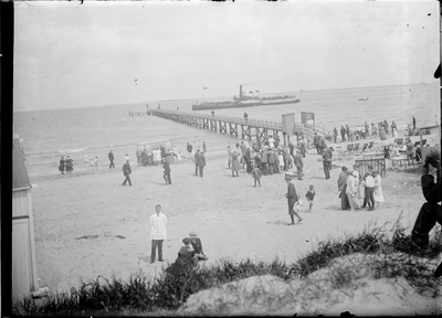GN2677 Kijkje op het strand met badgasten en de Noordzee. Langs het aanlegsteiger ligt de veerboot; ca. 1925