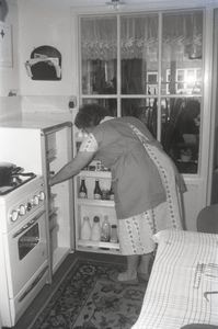 B1126 Een vrouw pakt iets uit de koelkast; ca. 1965