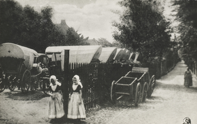 P-1390 Twee meisjes en een vrouw in Walcherse dracht te Middelburg. Op de achtergrond een stalling voor huifkarren aan ...