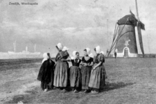 10008 Zeedijk Westkapelle met vrouwen en meisjes in klederdracht, en molen op de achtergrond (reproductie)