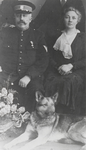 M 9013 Geüniformeerde man met pet, snor, onderscheiding op borst, ernaast een vrouw, aan hun voeten een herdershond.