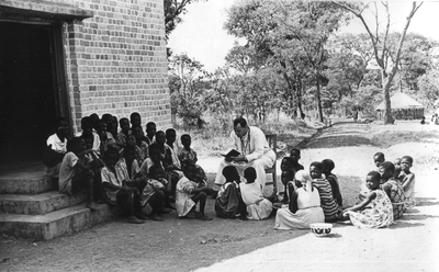 140084 Pater Ton Buijs leest in de buitenlucht voor aan de jeugd van de missiepost te Luwingu,Zambia