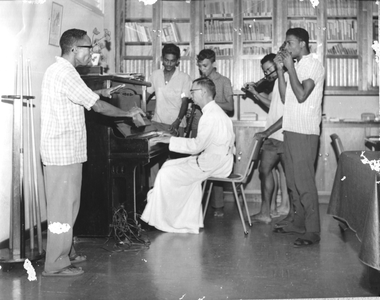186669 Rudy Wong Loi Sing dirigeert op het Petrus Donders seminarie te Paramaribo het orkest met pater Donicie aan de ...