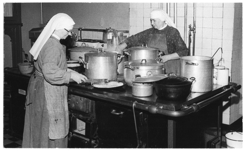 214118 Zuster Bernadette en zuster Imelda bereiden de warme maaltijd voor de communiteit in de keuken van het klooster ...