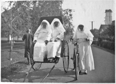 252286 Zuster Leontine met fiets naast een riksja met mère Isabelle en zuster Dorothea, in een straat te Medan op ...