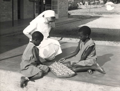 116075 Zuster-verpleegkundige met twee jonge patiënten voor een ziekenhuis te Zambia