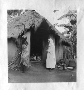 116135 Witte Zuster en Afrikaanse vrouw voor de deuropening van een huis, met binnen man en kinderen op de grond