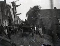 A1016 Feestelijke optocht met een paard met wagen en een muziekvaandel. Aan de gevels van de huizen hangen vlaggen.