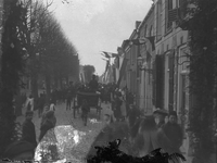 A1023 Feestelijke optocht met een paard met wagen en een muziekvaandel. Aan de gevels van de huizen hangen vlaggen.