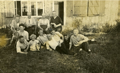 125 - Groepsfoto van elf mannen, één kind en een herdershond voor een barak