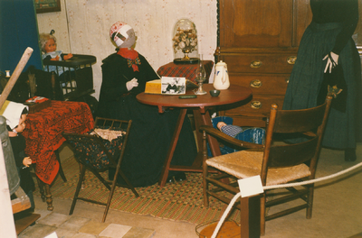 N 8126 - tafereeltje met pop in tipmutsdracht in stoel bij tafel met daarop een foto; opoe Grietje in de pratstoel; pop ...