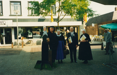 N 8132 - twee vrouwen, in dracht, een man in dracht en een in burgerpak, staan bij een pop die een vrouw in ...
