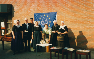 N 8133 - aantal mensen, twee in burger, de anderen in dracht, voor een muur met daarop een vlag met een logo; 5e open ...