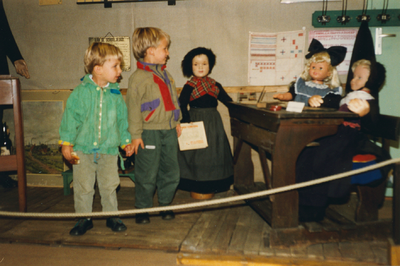 N 8138 - twee jongens bij een schoolbank met daarin en daarnaast poppen in oude dracht; sluiting oudheidkamer