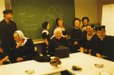 N 8143 - groep volwassenen en kinderen rond witte tafels; achtergrond schoolbord met wiskundeformule; vlnr: Rein ...