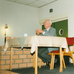 N 8150 - man, achter tafel met daarop beige kleed, vertelt een verhaal; Frank Hooghordel, lid streektaalgroep