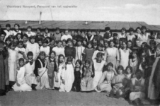 N 8820 - grote groep vrouwen en jonge meisjes, waarbij velen gekleed in witte werkkleding, staande voor een barak met ...