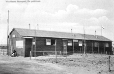 N 8832 - Een man zit aan de zijkant van een barak bij de paal van een Nuts-voorziening, één raam van de barak staat open.